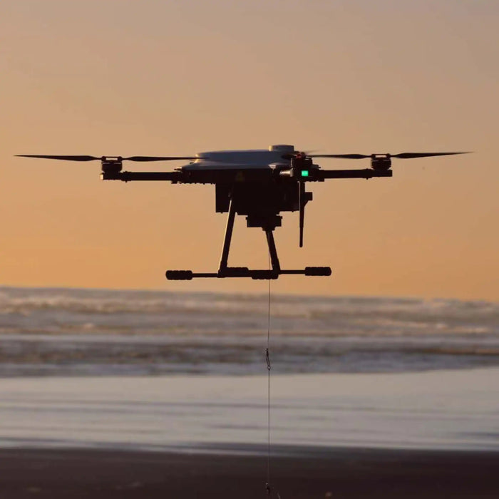 A black AEE Condor Pro Fishing drone flying over a beach at sunset, with its payload suspended by a thin cable below it.