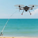 A black quadcopter drone hovering over a beach with the ocean in the background, holding a fishing rod extended downward. The drone has red accents on its landing legs and propeller guards.