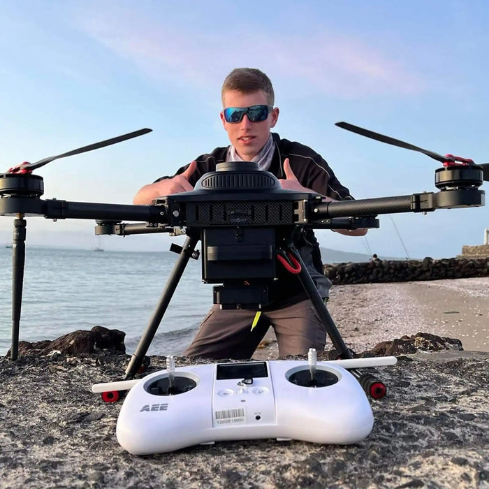 A man wearing sunglasses stands behind a large black Condor PRO fishing drone on a rocky beach, with a white remote control in the foreground and the ocean and sky behind him.
