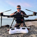 A man wearing sunglasses stands behind a large black Condor PRO fishing drone on a rocky beach, with a white remote control in the foreground and the ocean and sky behind him.