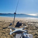 A white Condor A22S Fishing drone sits on a beach next to a fishing rod on a stand, with the ocean and shoreline in the background under a clear blue sky.