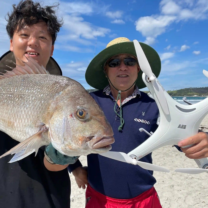 Two men on a beach holding a large fish and a white Condor A22S Fishing drone. One man wears a green hat, sunglasses, a navy polo shirt with 'Gucci' printed on it, and red shorts.