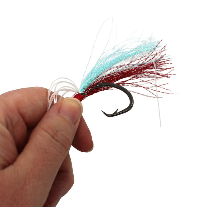 A close-up of a hand holding a fishing lure with a red body, black hook, white and light blue skirt strands, and red-and-white glitter fibers against a plain white background.