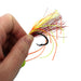 A close-up of a hand holding a fishing lure with a black hook and a skirt made of neon orange, yellow, red, and green strands on a white background.